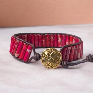 May include: A red and white stone bracelet with a gold heart-shaped button closure. The bracelet is made of leather and has a braided design.