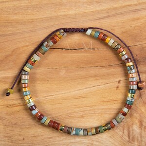 May include: A multi-colored beaded bracelet with a brown cord closure. The beads are made of natural stone and feature a variety of colors, including brown, red, green, and yellow.