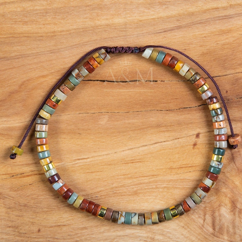 May include: A multi-colored beaded bracelet with a brown cord closure. The beads are made of natural stone and feature a variety of colors, including brown, red, green, and yellow.