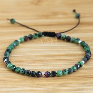 Puede incluir: Un brazalete de piedras preciosas verdes, negras y rosas con cuentas plateadas y cierre de cordón negro. El brazalete es ajustable y tiene un diseño sencillo.