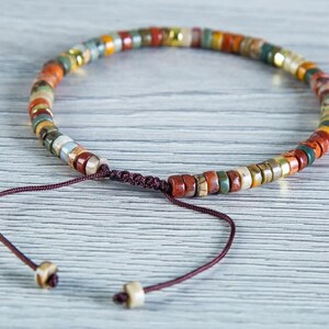 May include: A multi-colored beaded bracelet with a brown cord closure. The beads are made of natural stone and have a variety of colors, including red, orange, yellow, green, and brown.