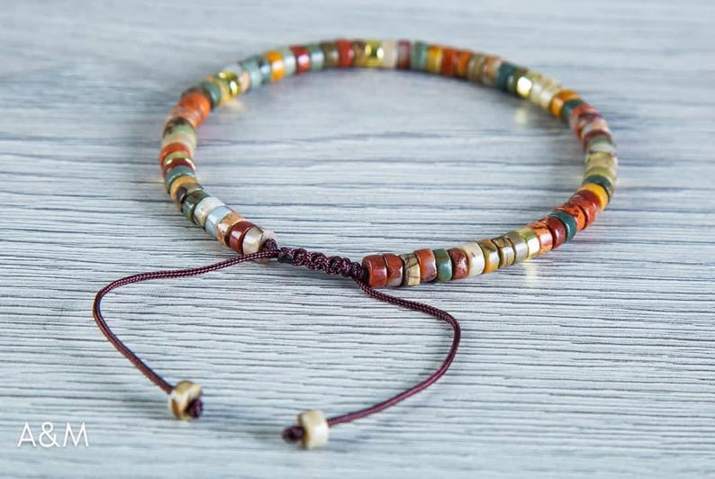 May include: A multi-colored beaded bracelet with a brown cord closure. The beads are made of natural stone and have a variety of colors, including red, orange, yellow, green, and brown.