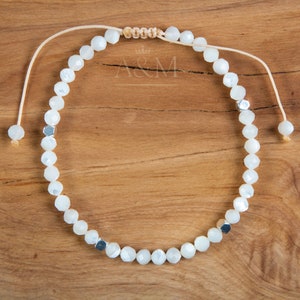 Puede incluir: Pulsera de cuentas blancas con detalles plateados y cierre de cordón beige. La pulsera está sobre una superficie de madera.