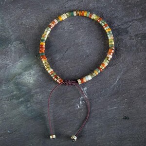May include: A multi-colored beaded bracelet with a brown cord and a sliding knot closure. The beads are small and round, and they are arranged in a random pattern. The bracelet is made of natural materials.