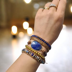 May include: A layered bracelet with a large oval lapis lazuli gemstone charm. The bracelet is made with brown leather cord, gold beads, and blue beads.