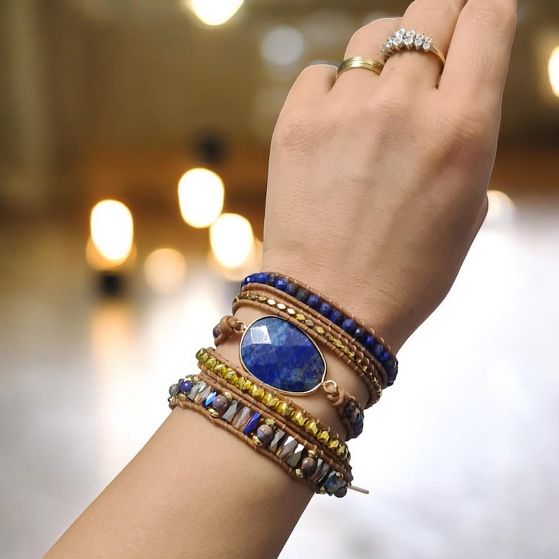 May include: A layered bracelet with a large oval lapis lazuli gemstone charm. The bracelet is made with brown leather cord, gold beads, and blue beads.