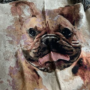 May include: A cream-colored woven throw blanket with a brown and black French Bulldog face printed on it. The dog's tongue is sticking out and its eyes are open.