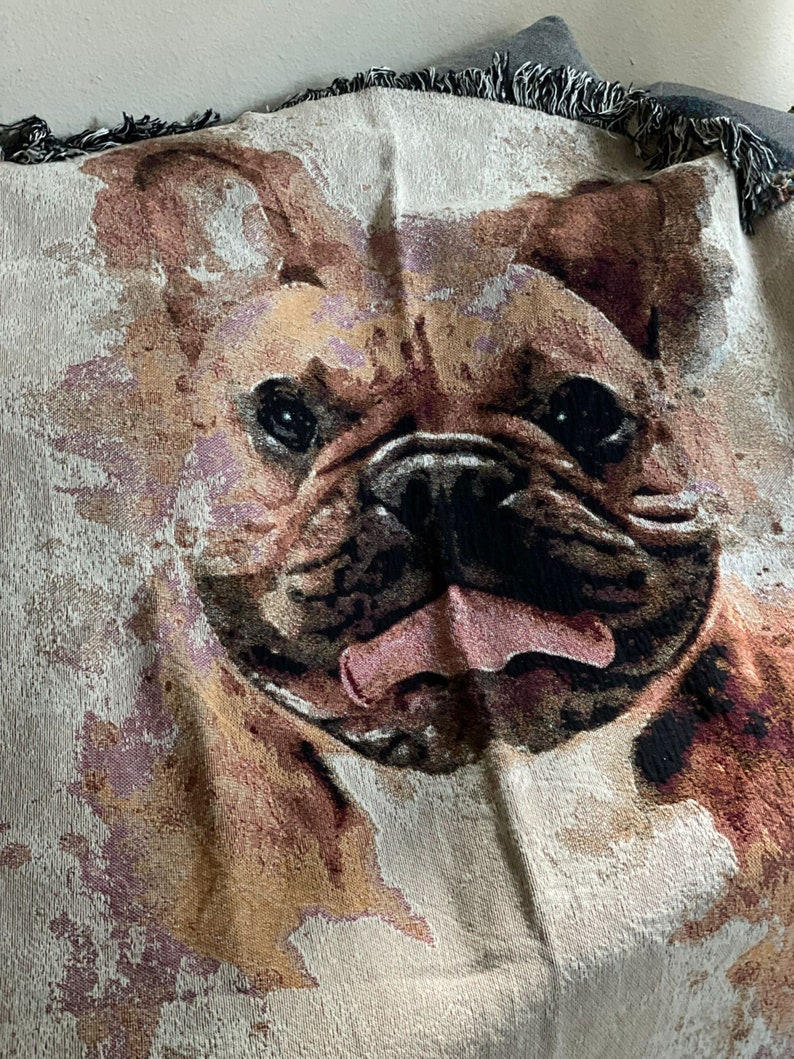 May include: A cream-colored woven throw blanket with a brown and black French Bulldog face printed on it. The dog's tongue is sticking out and its eyes are open.