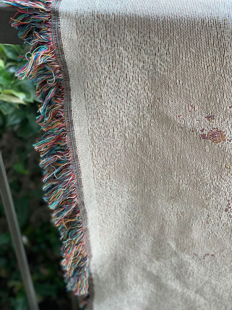 May include: A white woven throw blanket with a colorful fringe border. The blanket is made of a soft, textured material and has a subtle pattern.