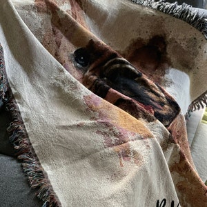 May include: A woven throw blanket with a brown and black dog face design. The blanket has a fringe border and the text "BK" is embroidered in black on the bottom right corner.