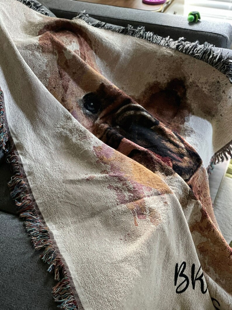 May include: A woven throw blanket with a brown and black dog face design. The blanket has a fringe border and the text "BK" is embroidered in black on the bottom right corner.