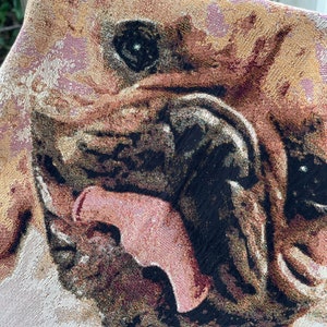 May include: Close-up of a tapestry featuring a brown and black dog with its mouth open and tongue sticking out. The dog's eyes are black and the background is a light beige color.