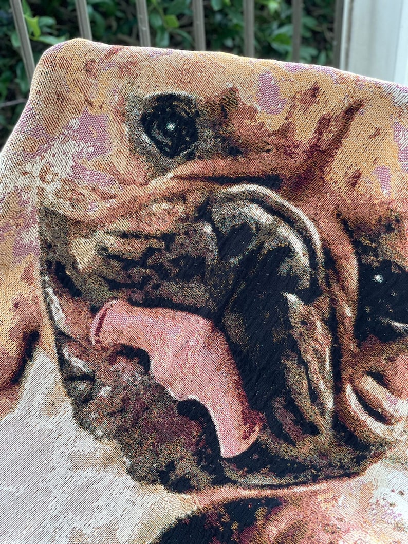 May include: Close-up of a tapestry featuring a brown and black dog with its mouth open and tongue sticking out. The dog's eyes are black and the background is a light beige color.