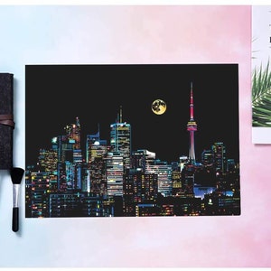 May include: A black canvas with a cityscape of Toronto, Canada, at night. The CN Tower is the tallest building in the image. The cityscape is illuminated with colorful lights. The canvas is ready for scratch art.