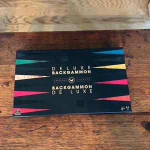 May include: A black box of the Deluxe Backgammon board game, with the text "DELUXE BACKGAMMON" and "BACKGAMMON DE LUXE" in gold lettering. The box has colorful accents and dice icons.