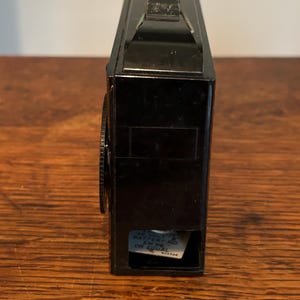 May include: A vintage black camera with a handle on top and a lens on the side. The camera has a rectangular shape and a small compartment on the bottom with text. The camera is sitting on a wooden surface.