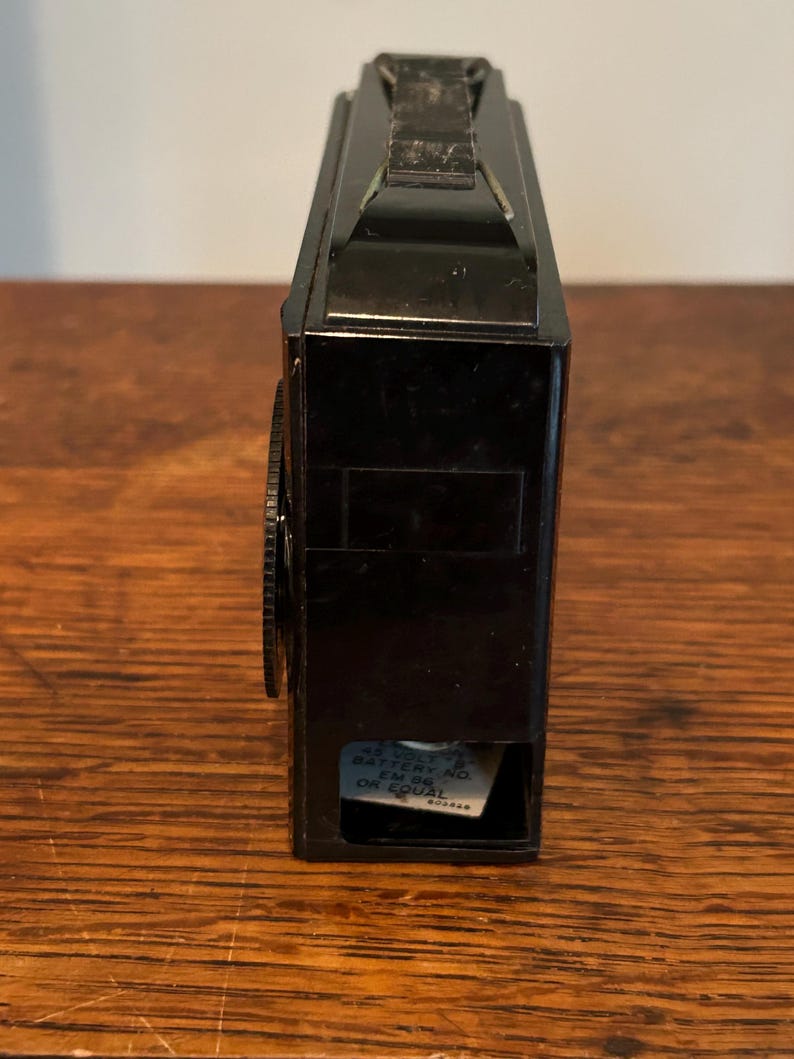 May include: A vintage black camera with a handle on top and a lens on the side. The camera has a rectangular shape and a small compartment on the bottom with text. The camera is sitting on a wooden surface.