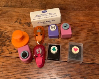 May include: A set of Creative Memories custom cutting system blade cartridges. The cartridges are in clear plastic cases and feature various shapes, including a butterfly, a music note, a footprint, a Christmas tree, and a heart. The cartridges are designed for use with the Creative Memories custom cutting system.