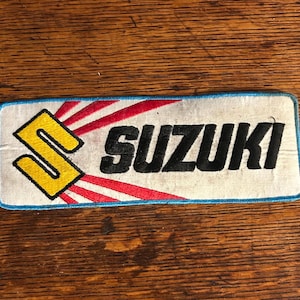 May include: Rectangular patch with the word "SUZUKI" in black, stylized font. The patch features a yellow "S" logo with a red and white sunburst design, all on a white background with a blue border.