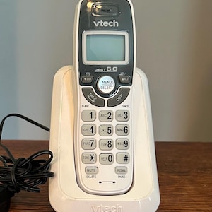 May include: A white and black Vtech cordless phone with a base. The phone has a screen and a keypad with numbers and function buttons. The base is white and has the Vtech logo.