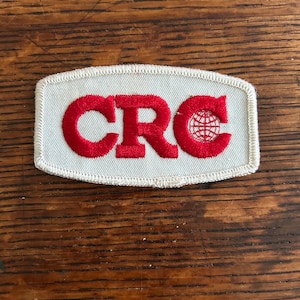 May include: Rectangular patch with the letters "CRC" in red, with a red globe graphic. The patch has a white background and a stitched border. The patch is on a wooden surface.