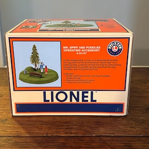 May include: A Lionel Mr. Spiff and Puddles Operating Accessory box. The box is orange and blue with a picture of a park scene. The scene includes a man, a dog, a woman, a bench, and a tree. The box also features the Lionel logo.