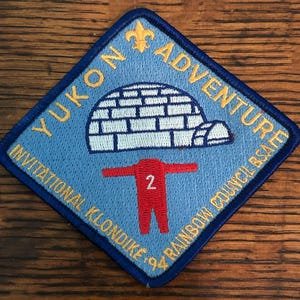 May include: A blue, diamond-shaped patch with gold text reading "YUKON ADVENTURE" and "INVITATIONAL KLONDIKE '94 RAINBOW COUNCIL BSA". The patch features a white igloo and a red figure with the number "2".