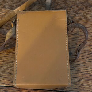 May include: A rectangular, tan leather case with a stitched border and a long, thin strap. The case has a smooth surface and appears to be vintage. The strap is a lighter shade of tan and is attached to the case with metal hardware.
