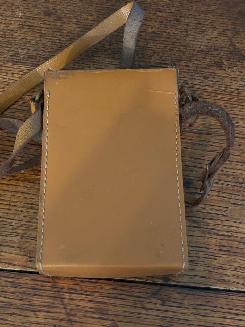 May include: A rectangular, tan leather case with a stitched border and a long, thin strap. The case has a smooth surface and appears to be vintage. The strap is a lighter shade of tan and is attached to the case with metal hardware.