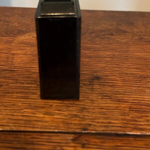 May include: A black rectangular object with a metallic top, possibly a vintage camera or a similar device. The object is positioned vertically, showcasing its sleek, dark surface. The background is a wooden surface.