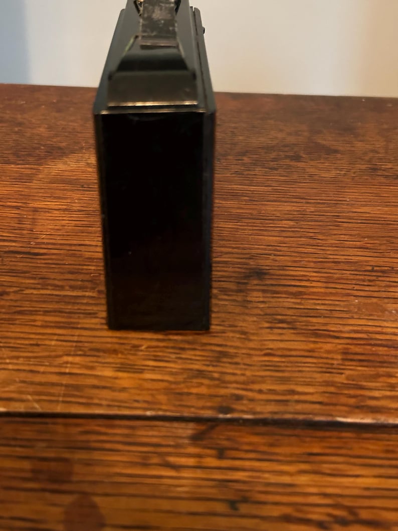 May include: A black rectangular object with a metallic top, possibly a vintage camera or a similar device. The object is positioned vertically, showcasing its sleek, dark surface. The background is a wooden surface.