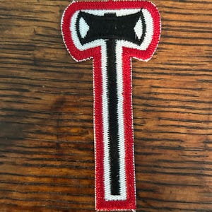 May include: Embroidered patch featuring a black axe head design with white and red borders. The axe head is centered above a long, vertical handle, also in black, white, and red. The patch is likely for clothing or accessories.