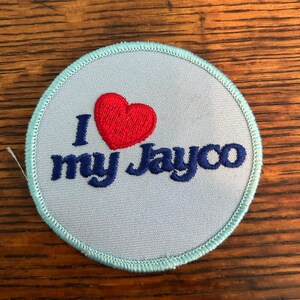 May include: A round patch with a light blue background and a teal border. The patch features the text "I my Jayco" in dark blue, with a red heart above the word "my".