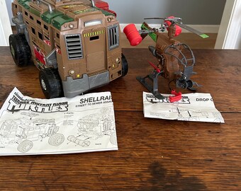 May include: Teenage Mutant Ninja Turtles Shellraiser vehicle and helicopter playset. The Shellraiser is a brown, green, and gray vehicle with a red cannon on top. The helicopter is brown and green with red accents. The playset comes with instructions for assembly.