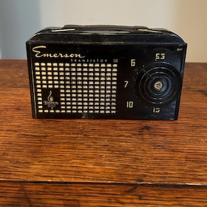 May include: A vintage black Emerson Transistor II radio with a handle. The radio features a speaker grill, a tuning dial, and the numbers 6, 7, 10, and 15. The radio is sitting on a wooden surface.