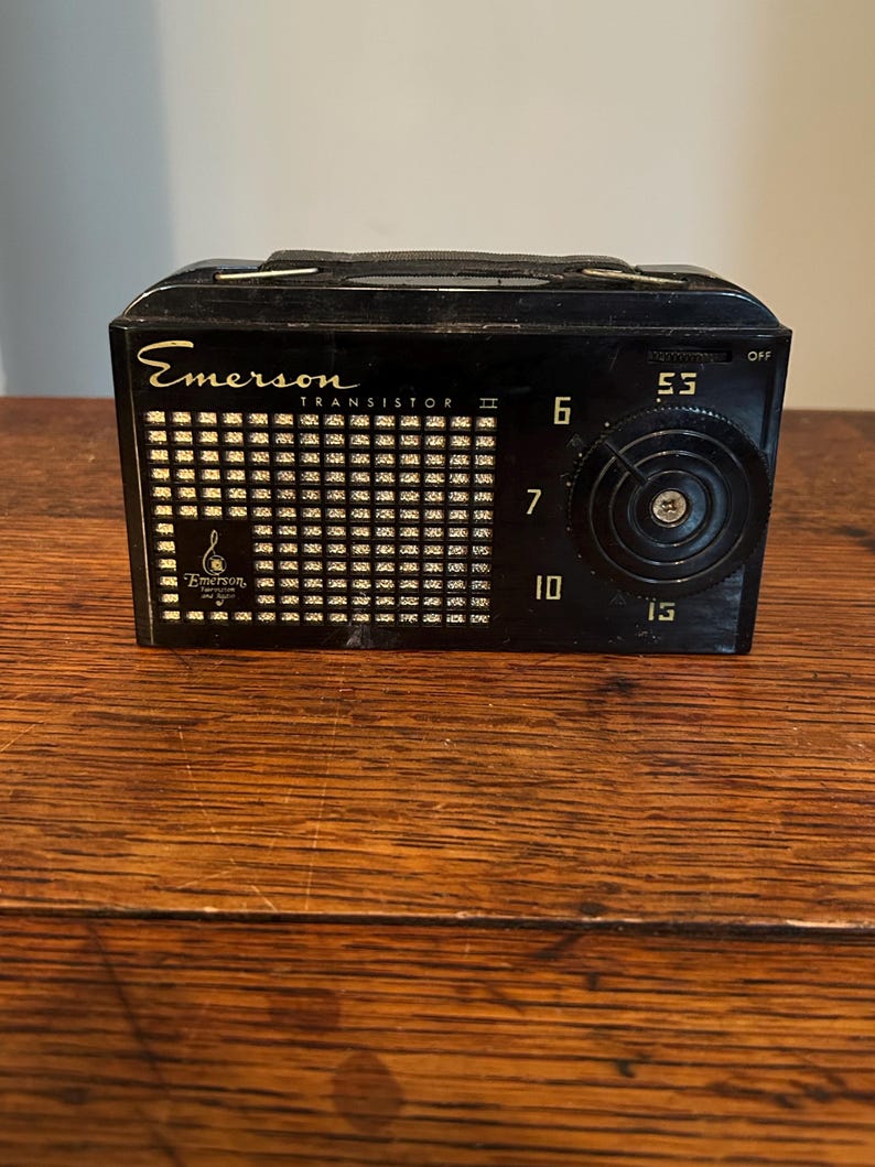 May include: A vintage black Emerson Transistor II radio with a handle. The radio features a speaker grill, a tuning dial, and the numbers 6, 7, 10, and 15. The radio is sitting on a wooden surface.