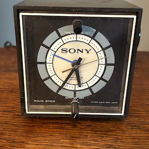 May include: A vintage black Sony alarm clock with a white and blue face. The clock face has a circular design with the brand name "SONY" in the center. It features a blue second hand and a black control knob. The text "SOLID STATE" and "CLOCK U.S.A. - DIAL JAPAN" are visible.