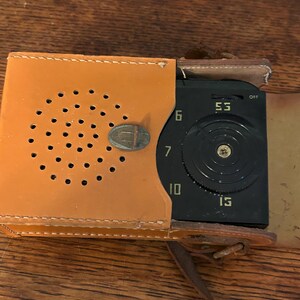May include: Vintage portable radio in a tan leather case. The radio has a black face with a tuning dial and the numbers 53, 6, 7, 10, and 15. The case has a series of small holes and a metal clasp.