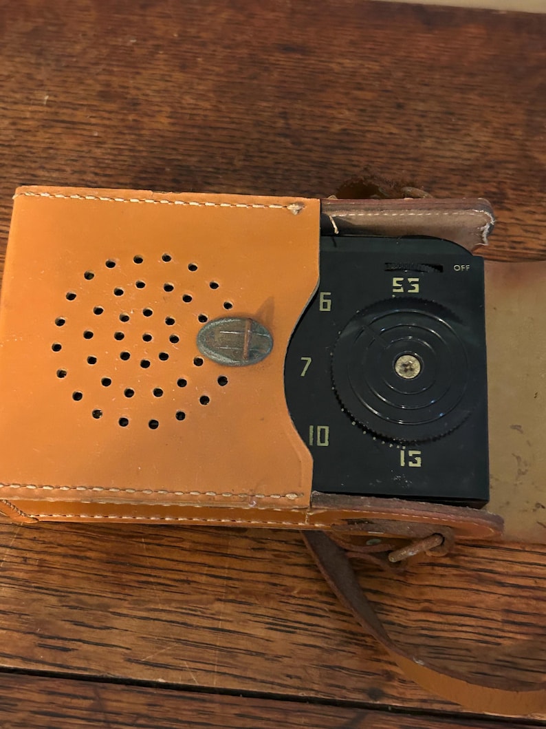 May include: Vintage portable radio in a tan leather case. The radio has a black face with a tuning dial and the numbers 53, 6, 7, 10, and 15. The case has a series of small holes and a metal clasp.
