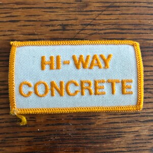 May include: A rectangular patch with the words "HI-WAY CONCRETE" embroidered in orange thread on a white background. The patch has a yellow border and is likely a uniform or workwear accessory.