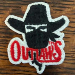 May include: Embroidered patch featuring a masked cowboy in a black hat and bandana. The word "Outlaws" is written in red, outlined in white. The patch is likely for clothing or accessories.