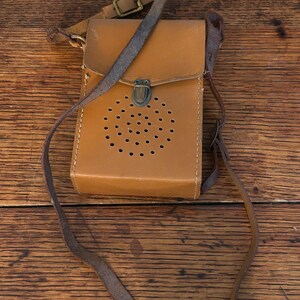 May include: A vintage tan leather crossbody bag with a flap closure and a series of small circular perforations. The bag features a long, thin strap and a smaller strap with a buckle. The metal clasp is a dark bronze color.