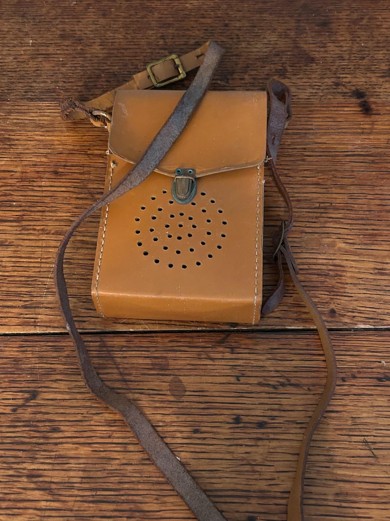 May include: A vintage tan leather crossbody bag with a flap closure and a series of small circular perforations. The bag features a long, thin strap and a smaller strap with a buckle. The metal clasp is a dark bronze color.