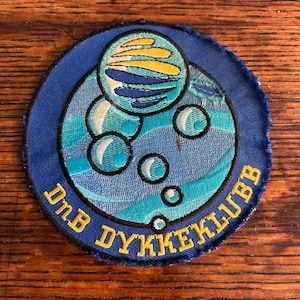 May include: A circular embroidered patch with a blue border featuring a central graphic of a stylized planet or sphere with yellow and blue stripes above light blue water bubbles. The text DnB DYKKEKLUBB is stitched in yellow along the bottom edge.
