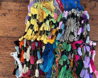 May include: A collection of colorful embroidery floss in various shades, including yellow, blue, green, pink, and purple. The floss is neatly bundled and arranged on a wooden surface, ideal for crafting and embroidery projects.