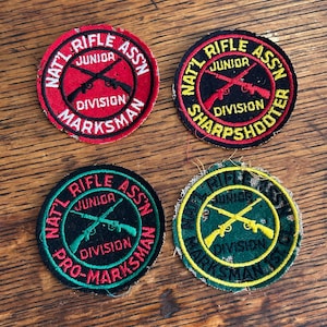 May include: Four vintage circular patches with text and crossed rifles. The patches are red, black, green, and yellow, with text that reads "Nat'l Rifle Assn Junior Division Marksman", "Sharpshooter", and "Pro-Marksman".