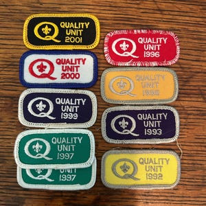 May include: A collection of rectangular patches with the text "QUALITY UNIT" and various years embroidered on them. The patches feature a stylized fleur-de-lis and the letter "Q". Colors include black, red, blue, green, and yellow.