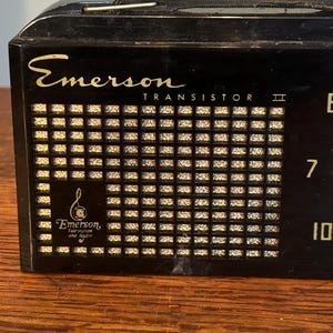 May include: Black Emerson Transistor II radio with gold lettering and a speaker grill. The radio has a tuning dial and the Emerson logo. The radio is sitting on a wooden surface.