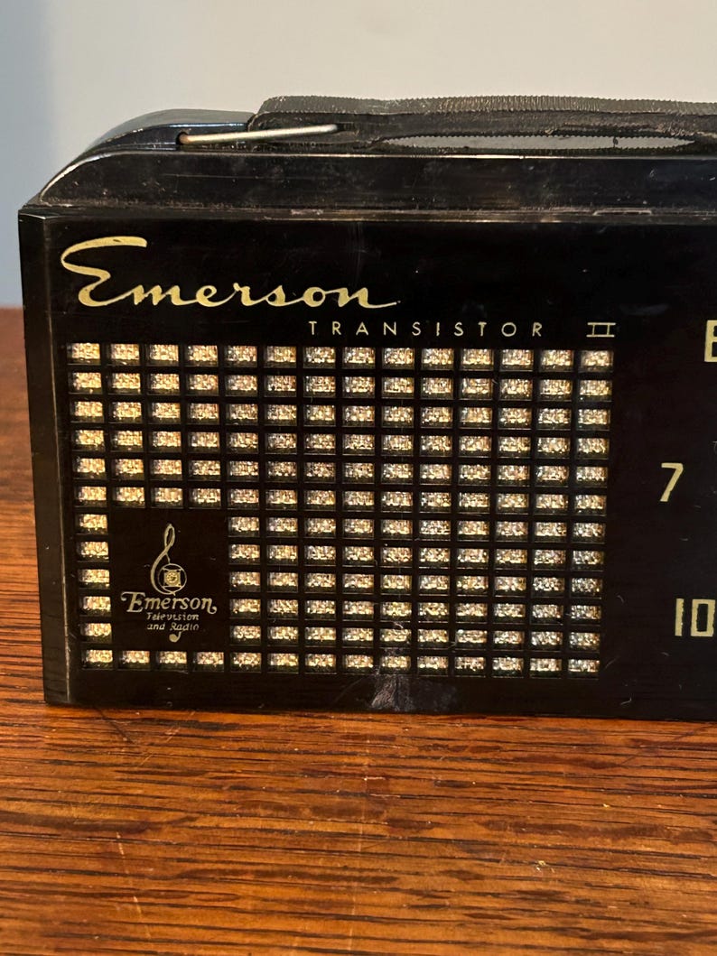 May include: Black Emerson Transistor II radio with gold lettering and a speaker grill. The radio has a tuning dial and the Emerson logo. The radio is sitting on a wooden surface.
