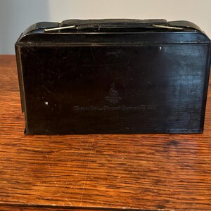 May include: A black, rectangular case with a handle and a clasp. The case has a glossy finish and appears to be made of a hard material. The text on the case is blurry.